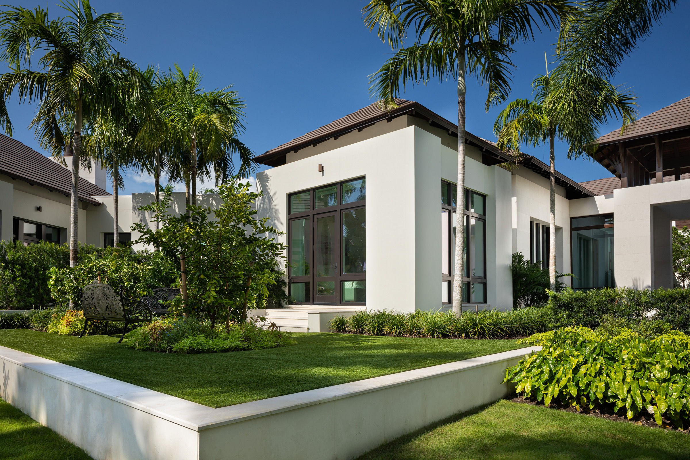 Modern white house surrounded by lush greenery and palm trees under a clear blue sky, featuring large windows and a manicured lawn.