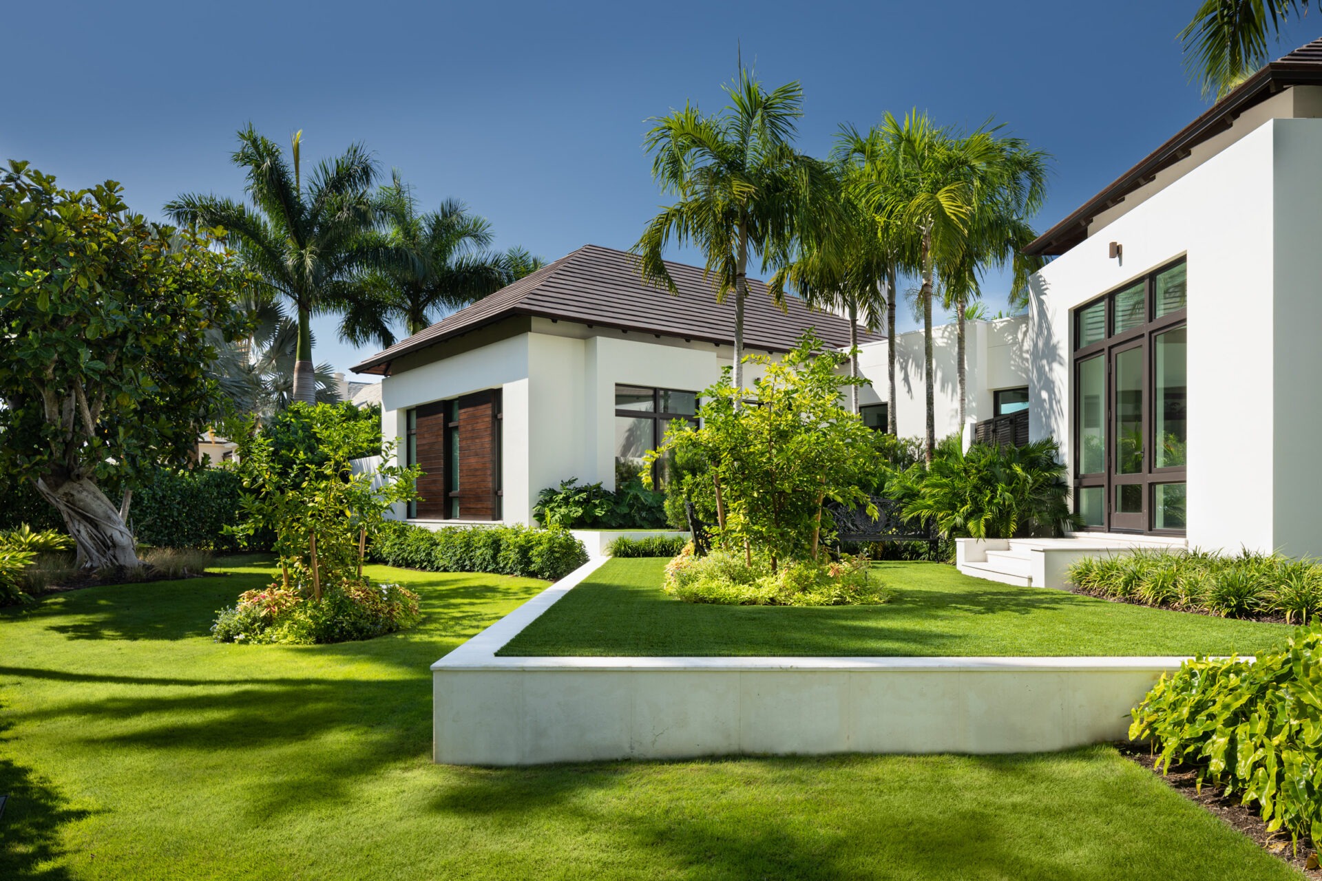 Modern white house with large windows, surrounded by lush green landscaping and palm trees under a clear blue sky. Peaceful and sunny atmosphere.