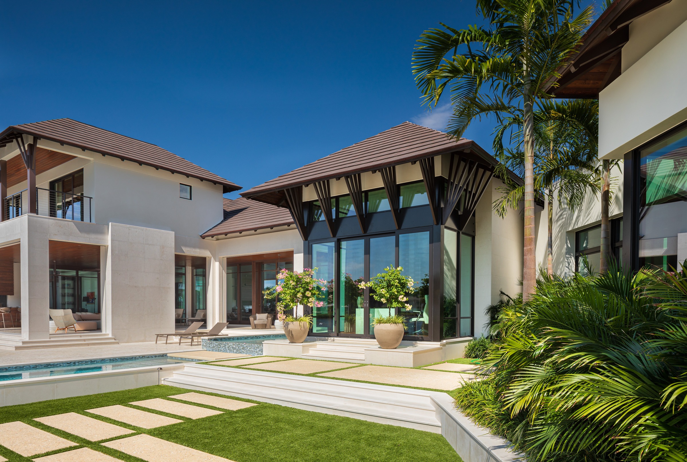 Modern luxury villa with large windows, tropical landscaping, and a pool. Clear blue sky and palm trees enhance the serene ambiance.