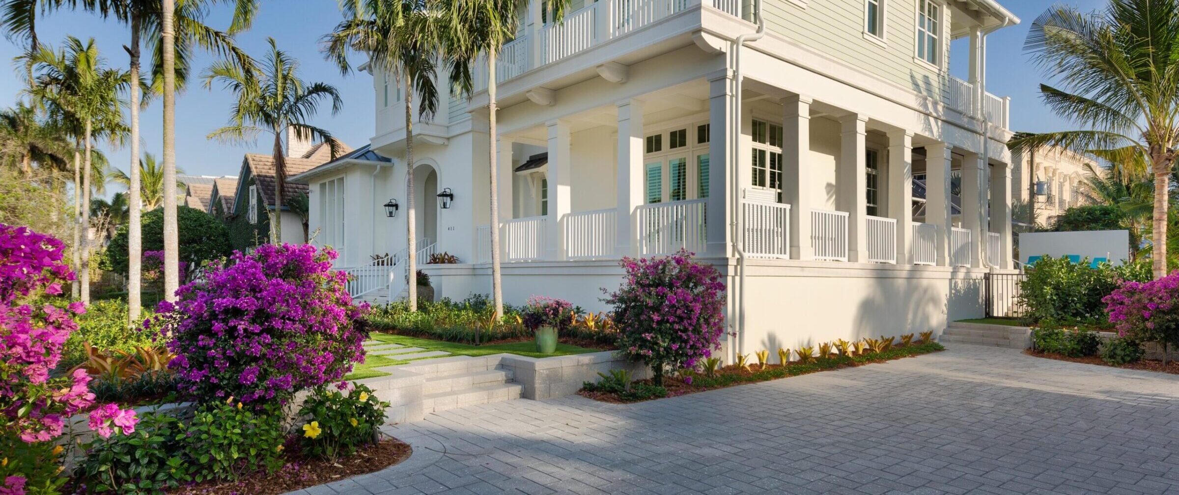 Tropical house with palm trees and vibrant flowers in the foreground, featuring a white porch and lush landscaping under a clear blue sky.