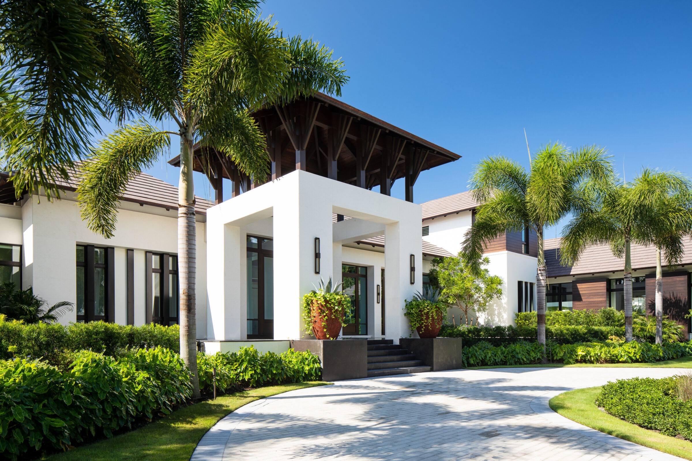 Modern house with tropical landscaping, featuring palm trees and lush greenery, set against a clear blue sky, with a welcoming front entrance.