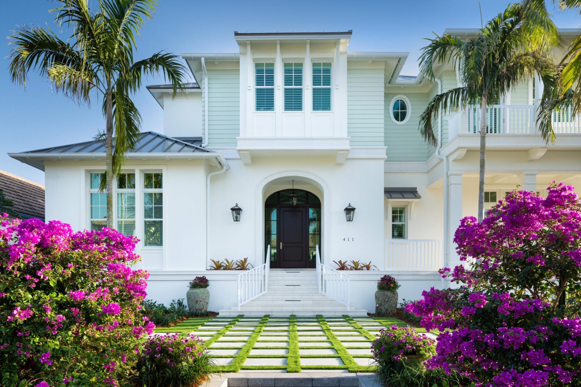 Elegant two-story white house with blue accents, surrounded by vibrant purple flowers and palm trees. Neat pathway leads to dark wooden door.