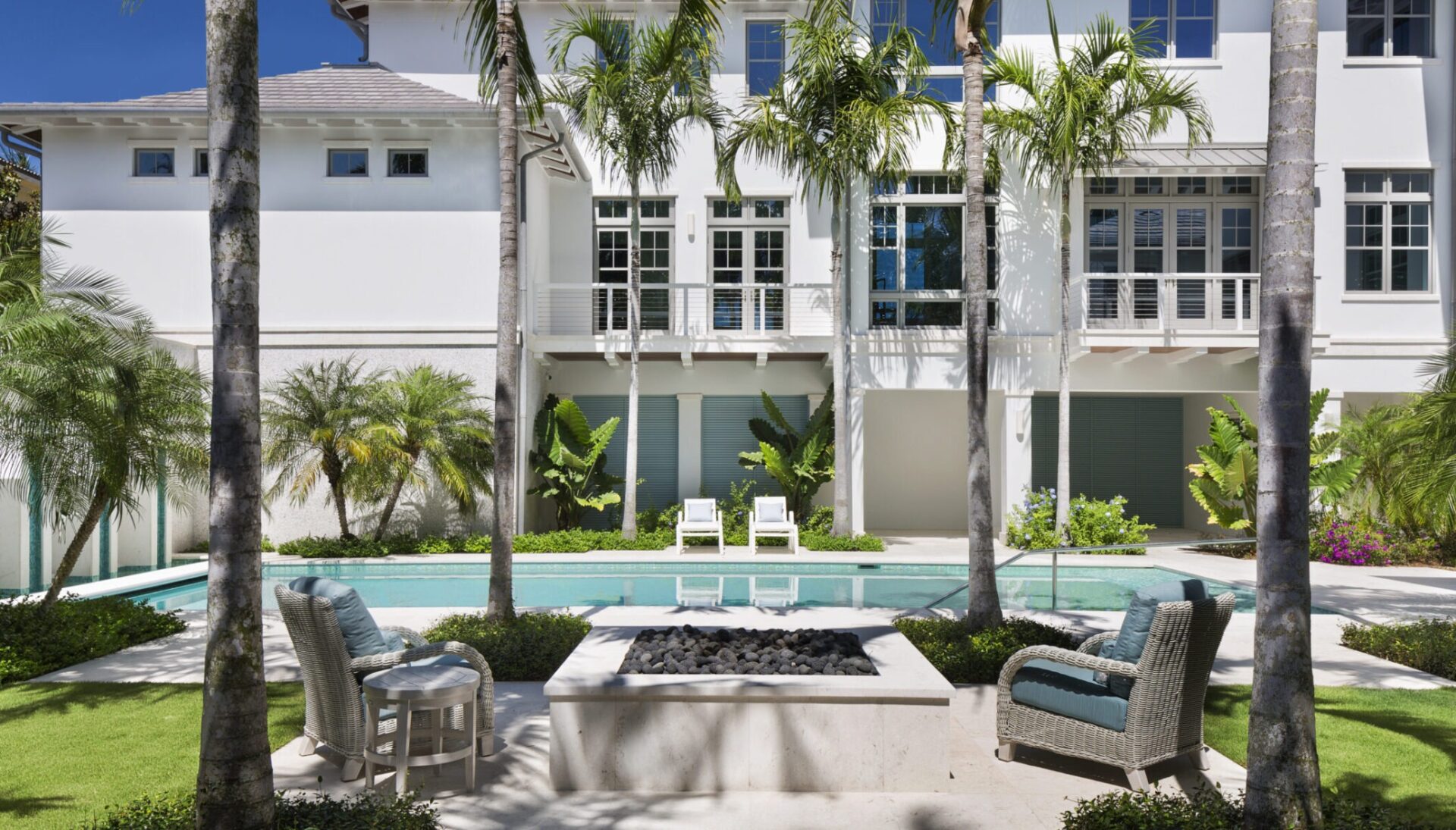 Modern white house with large windows, surrounded by palm trees and lush greenery. A pool and patio area with seating in the foreground.