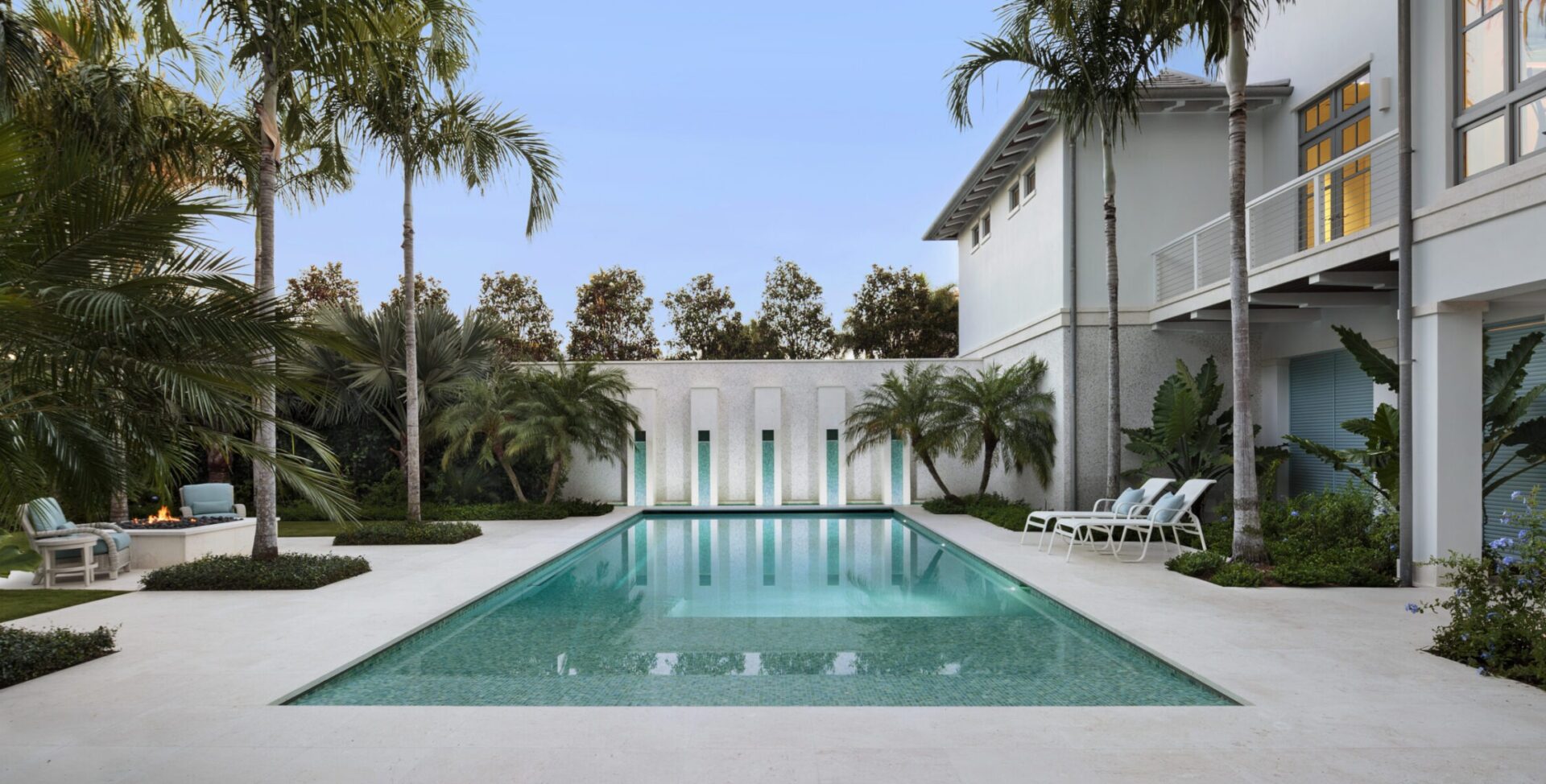 Luxurious patio with a rectangular pool, surrounded by palm trees and a modern building. Lounge chairs and greenery create a serene atmosphere.