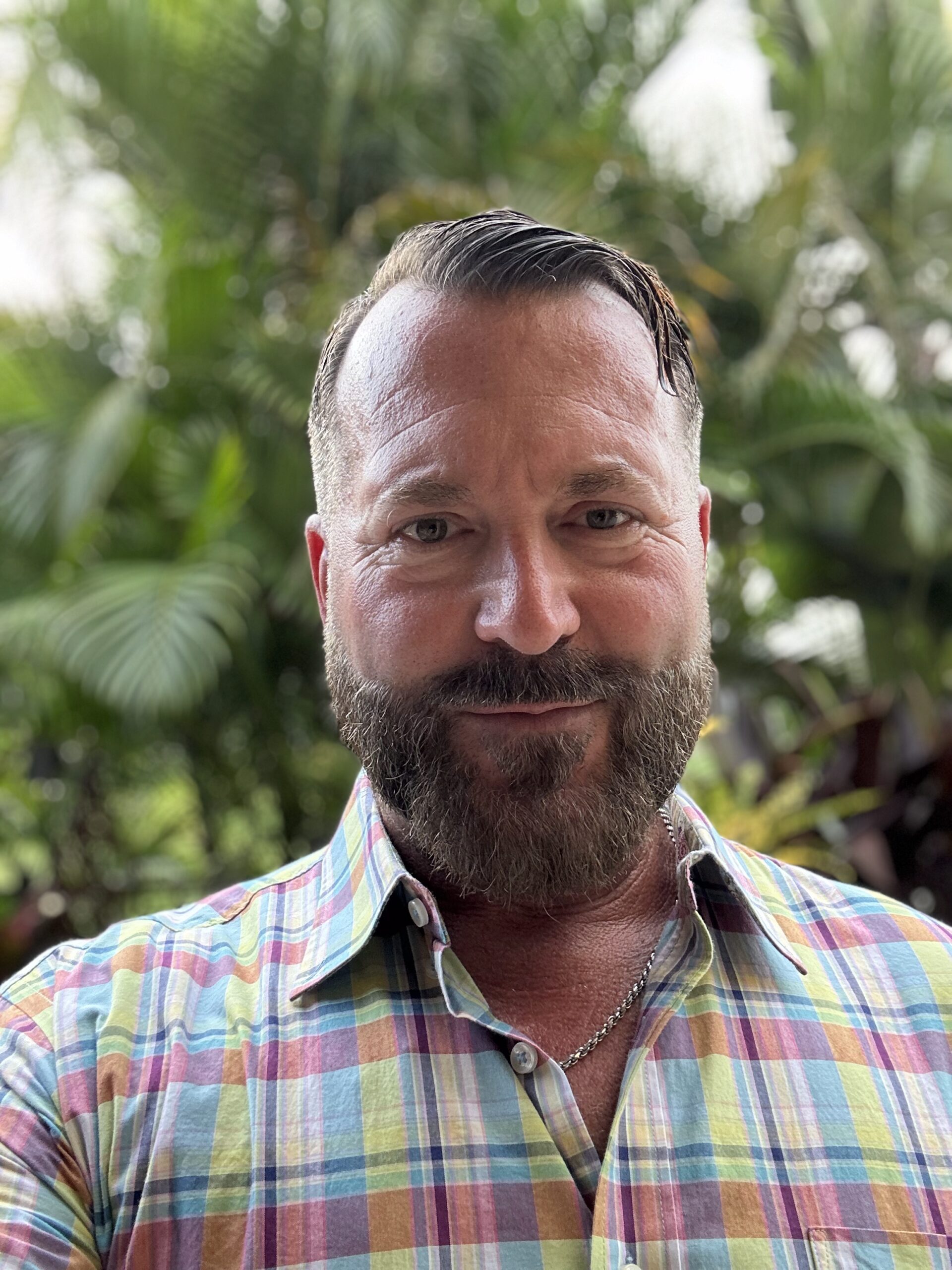 A person with a beard, wearing a colorful plaid shirt, stands outdoors with lush green foliage in the blurred background.