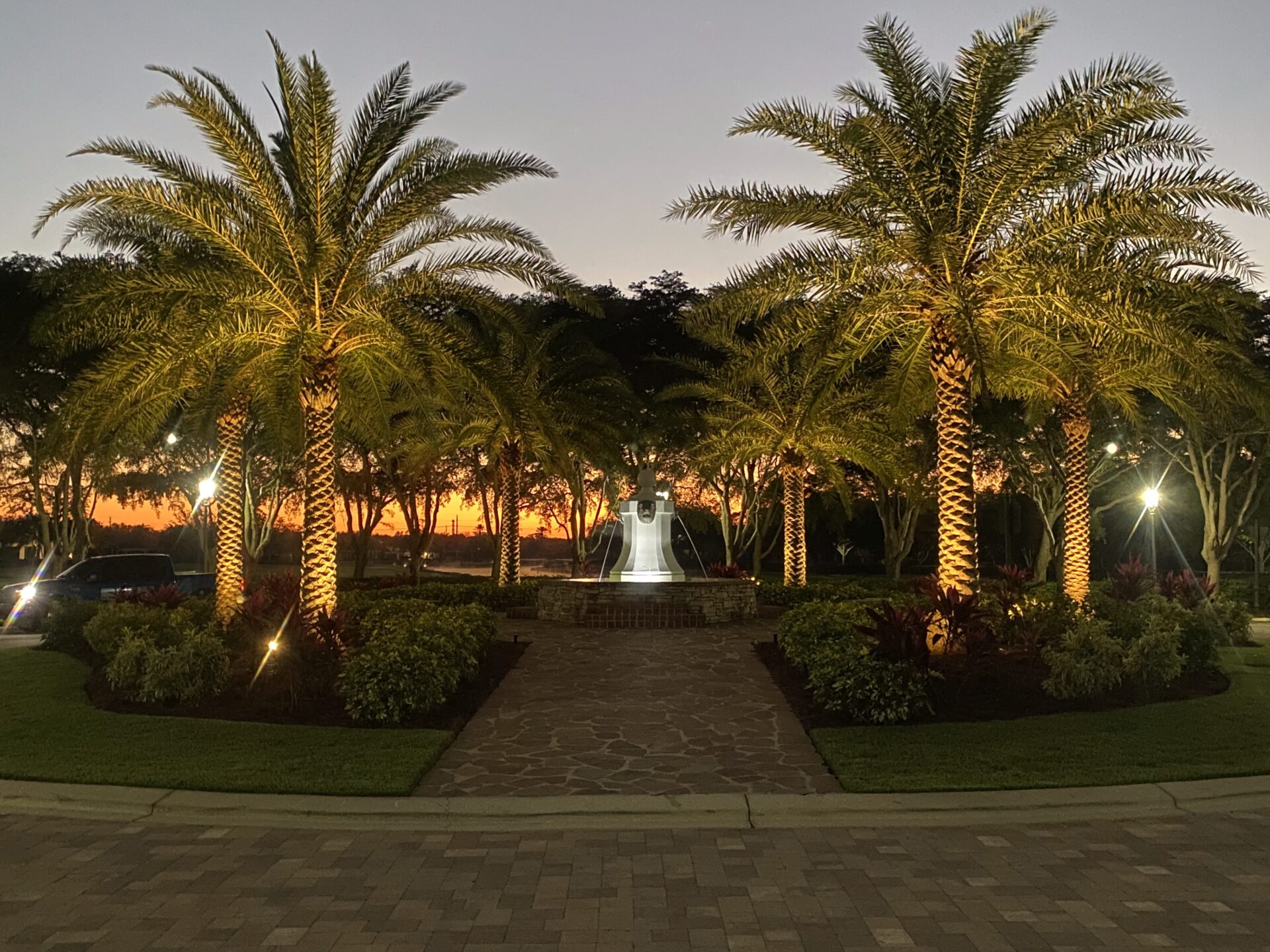 Palm trees line a path with illuminated fountain, surrounded by lush greenery against a sunset sky, creating a tranquil evening scene.