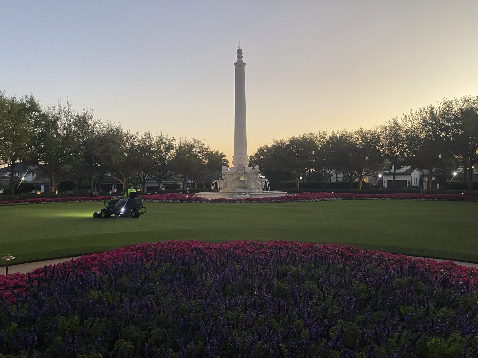 A person on a riding mower tends grass near the San Jacinto Monument, surrounded by blooming flowers and trees during dawn.