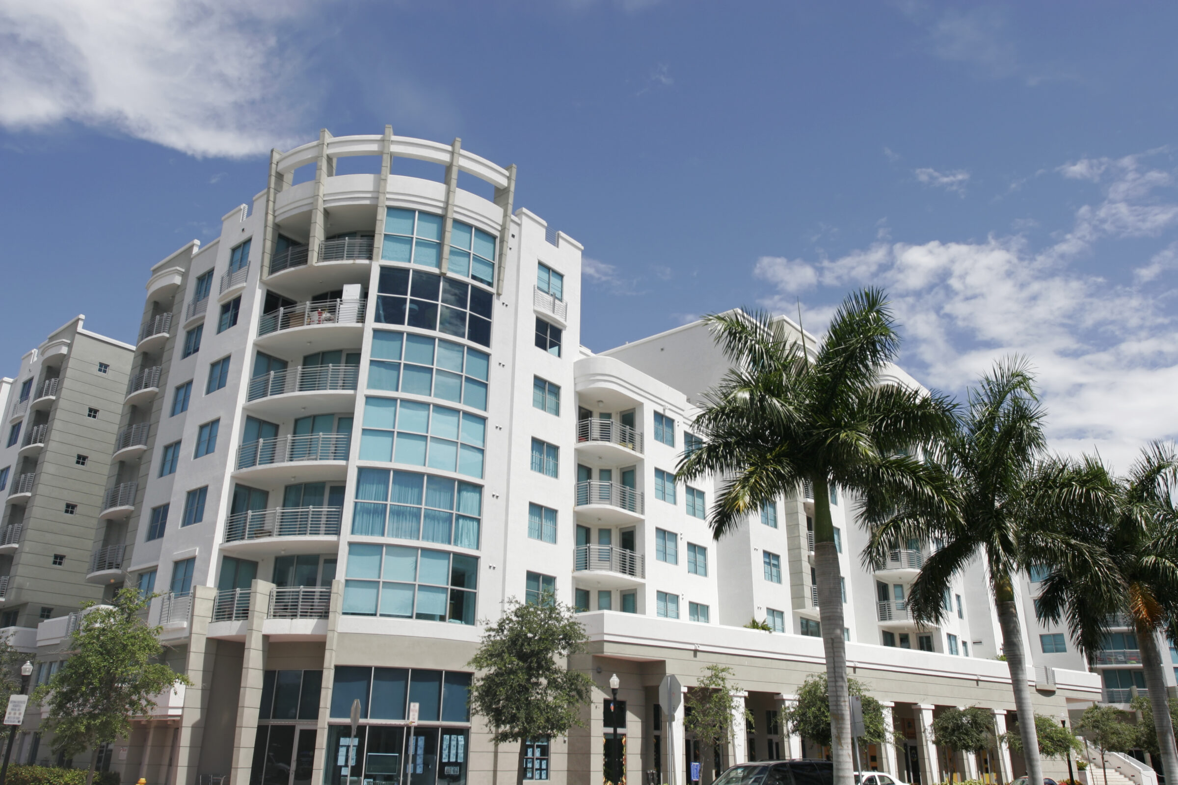 Modern urban apartment building with curved balconies and many windows, surrounded by lush palm trees under a clear blue sky.