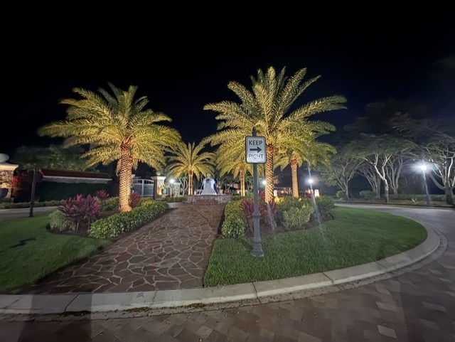 A nighttime pathway lined with illuminated palm trees leads to a fountain, surrounded by landscaped greenery and lit by streetlamps.
