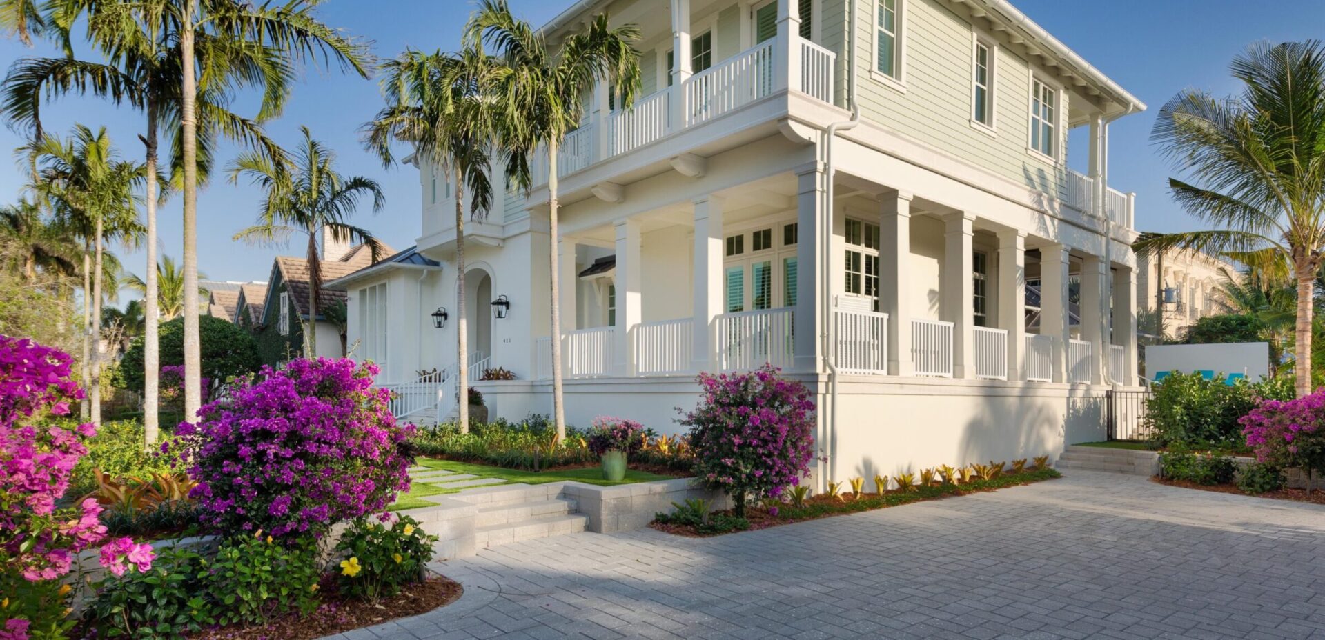 Colonial-style house with a spacious porch, surrounded by vibrant bougainvillea, palm trees, and a neatly paved driveway under a clear blue sky.