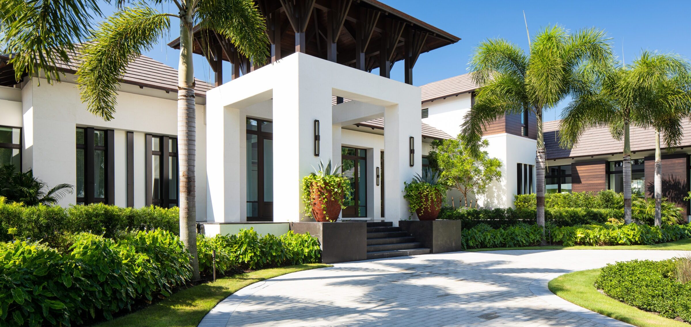 Modern house with white walls, large windows, surrounded by lush greenery, palm trees, and a circular driveway under a clear blue sky.