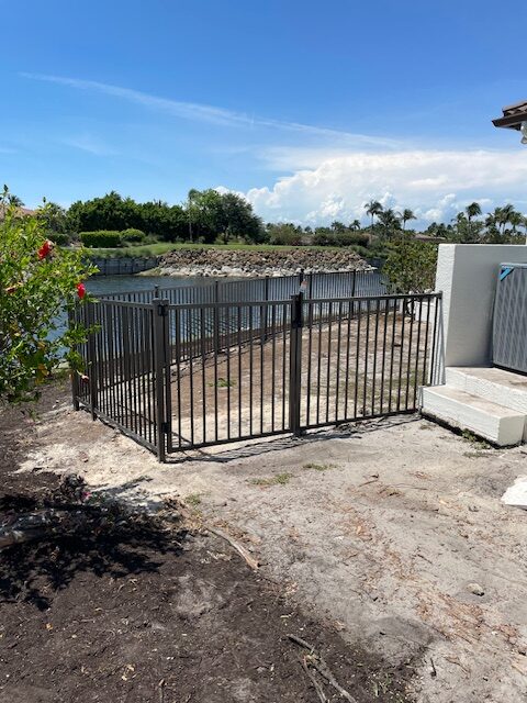 A fenced area near a waterway with a metal gate, surrounded by greenery, under a clear blue sky, adjacent to a building corner.