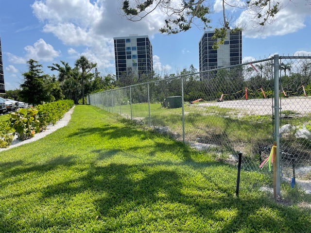 Fenced grassy pathway beside a vacant lot under construction, with three tall buildings in the background and trees lining the sidewalk.