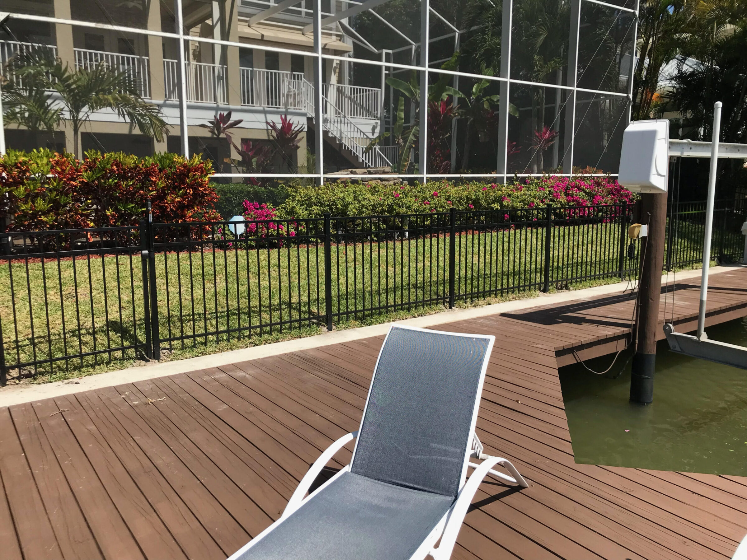 Lounge chair on wooden deck by water, black fence, landscaped garden, and a screened-in structure with white railings and tropical plants.