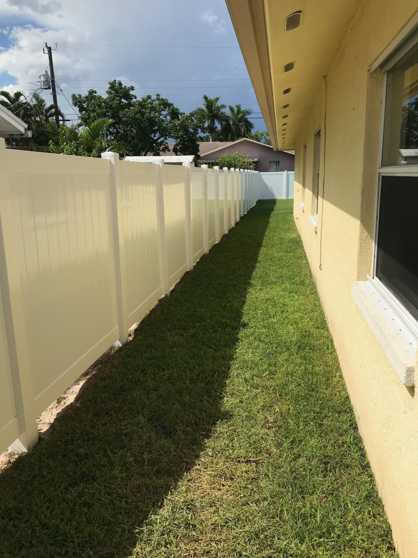 Narrow grassy path between a house and white fence, surrounded by trees and clear skies under sunlight. No people or landmarks visible.